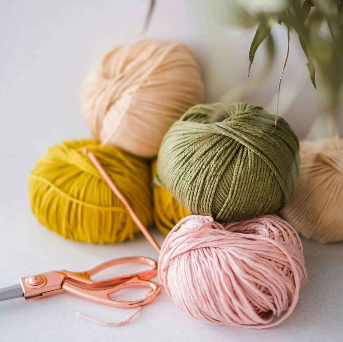Four balls of yarn in beige, yellow, green, and pink with a pair of scissors on a light background.