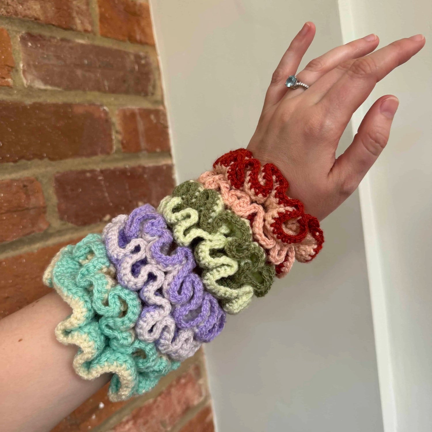 Colorful crocheted scrunchies on a wrist with a brick wall background