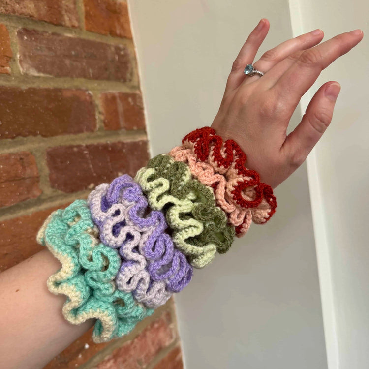 Colorful crocheted scrunchies on a wrist with a brick wall background