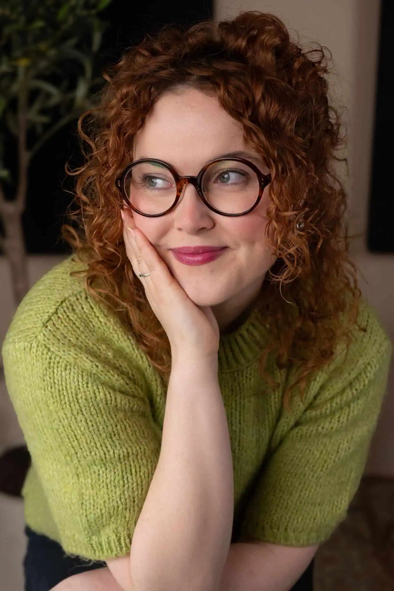 Woman with curly red hair wearing glasses and a green sweater, posing thoughtfully.