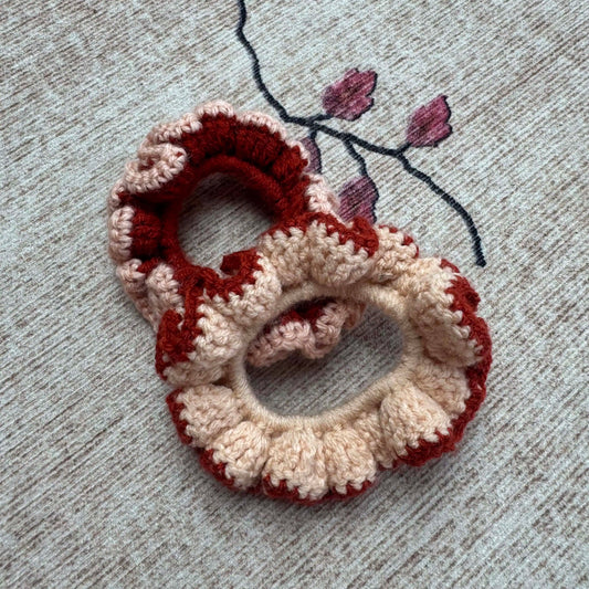 Two crocheted scrunchies in red and beige on a textured surface with floral embroidery.