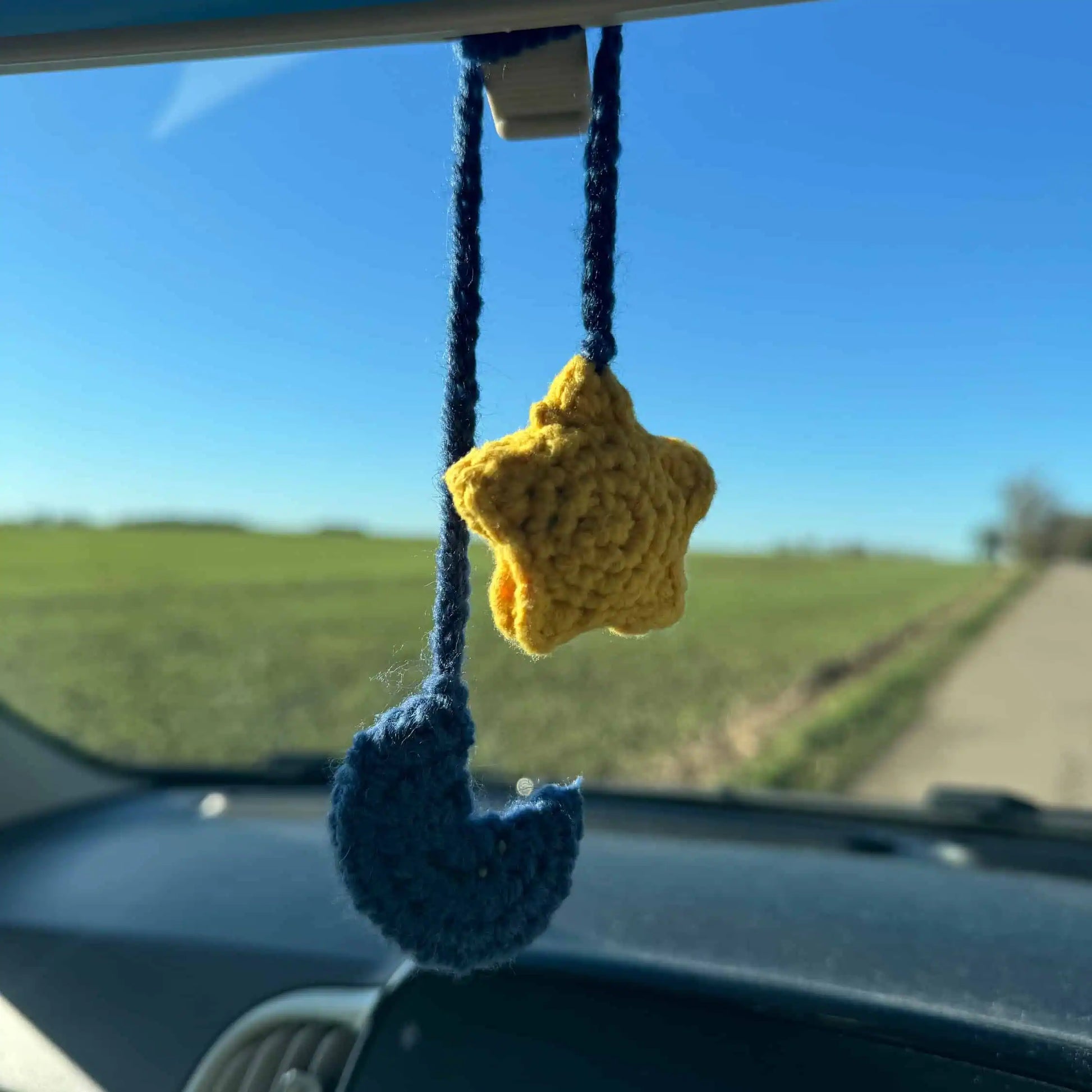 Crocheted star and moon hanging from a car's rearview mirror with a road and field view outside.