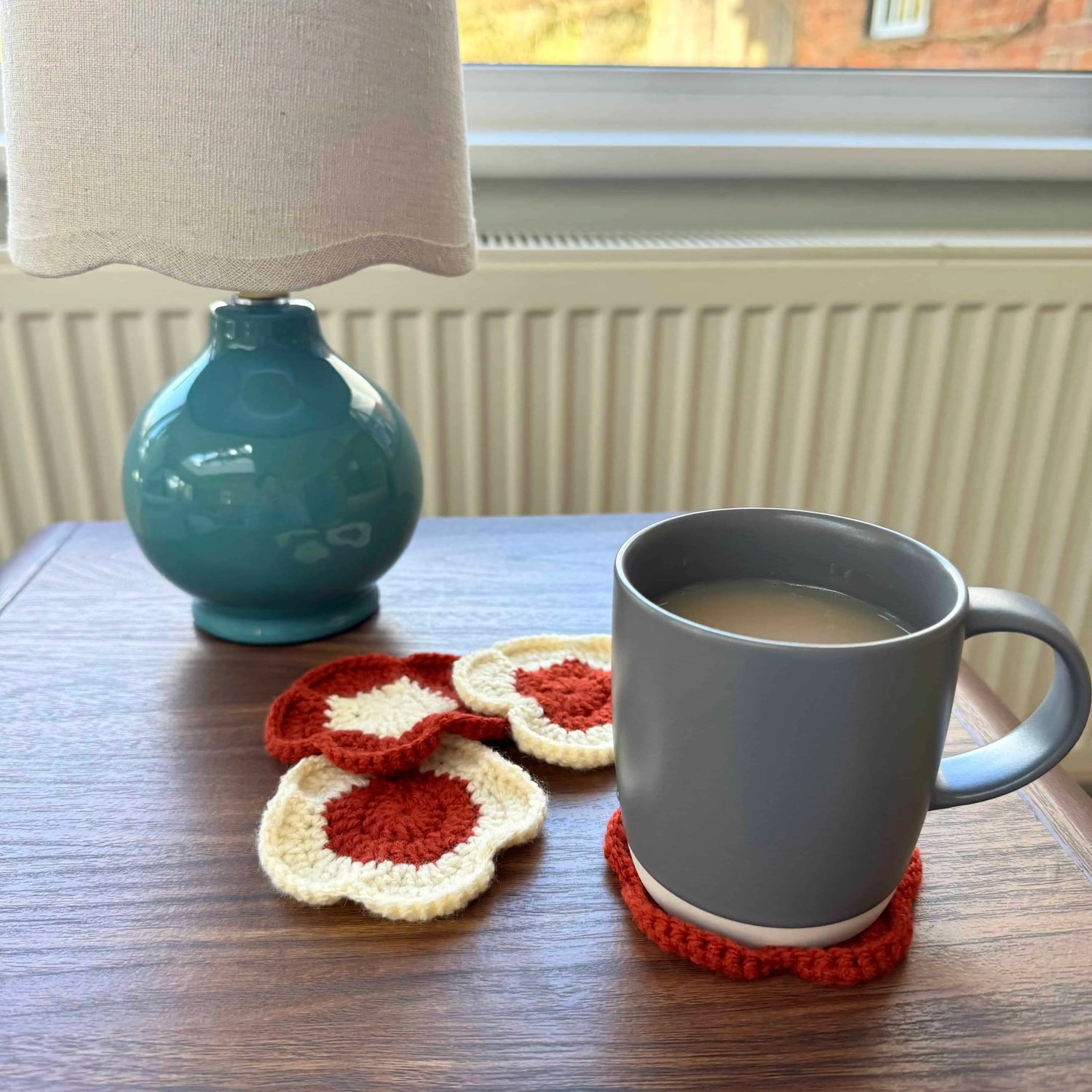Grey mug on a wooden table with colourful coasters, next to a blue lamp.