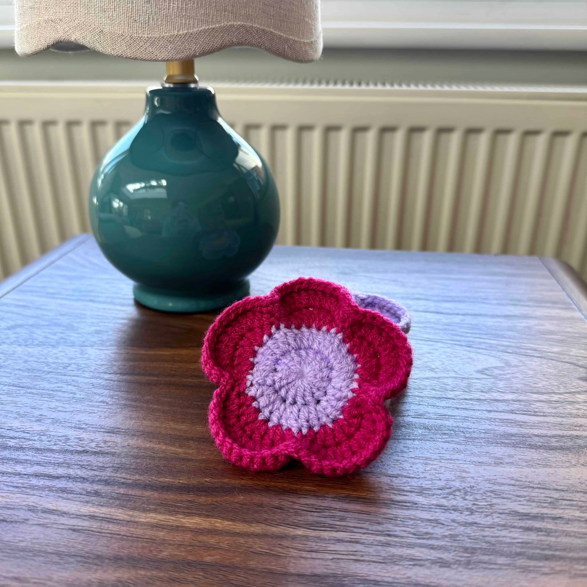 Crocheted coaster on a wooden surface next to a teal lamp.