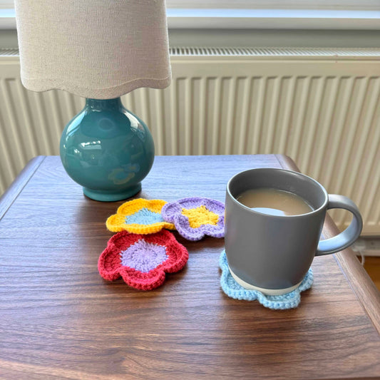 Gray mug with a floral coaster on a wooden table next to a lamp and colorful coasters.