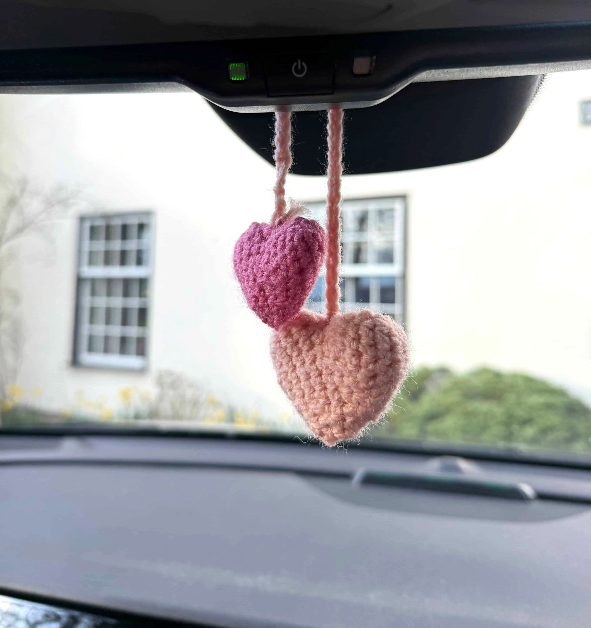 Two pink crocheted hearts hanging from a car's rearview mirror with a blurred outdoor background.