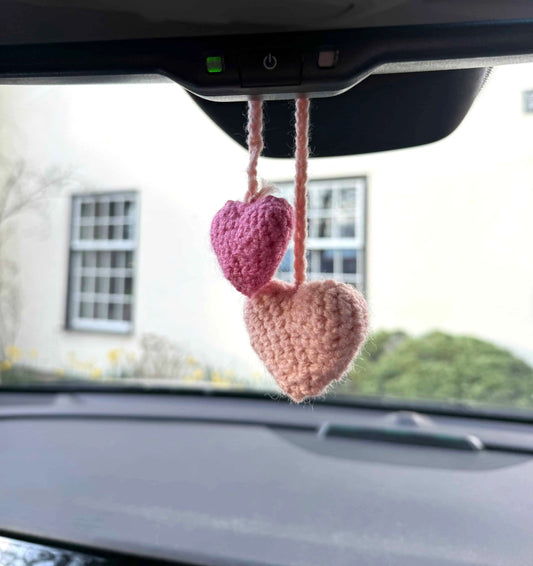 Two pink crocheted hearts hanging from a car's rearview mirror with a blurred outdoor background.