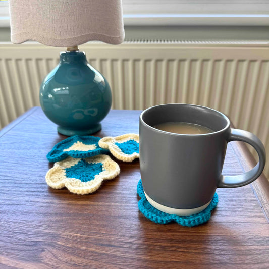 Gray mug with a blue crochet coaster on a wooden table next to a teal lamp.