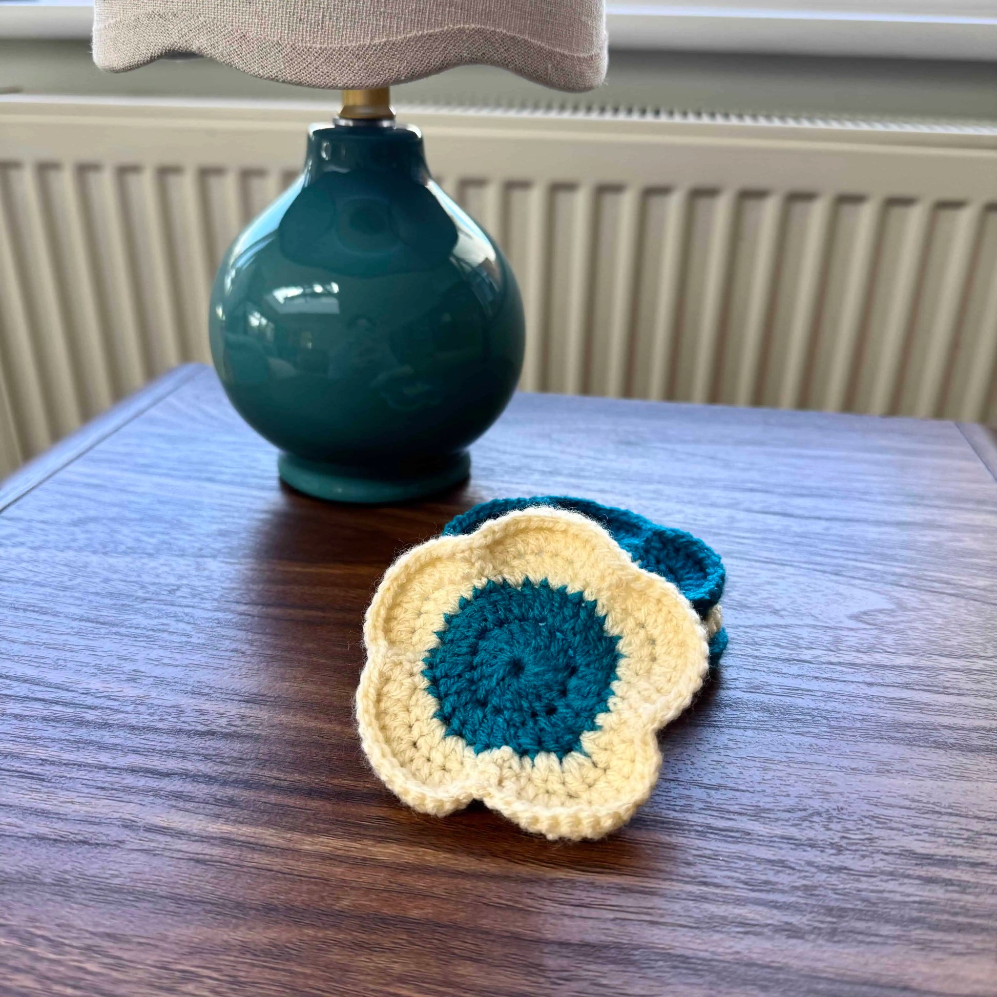 Crocheted coaster on a wooden surface with a teal lamp in the background.