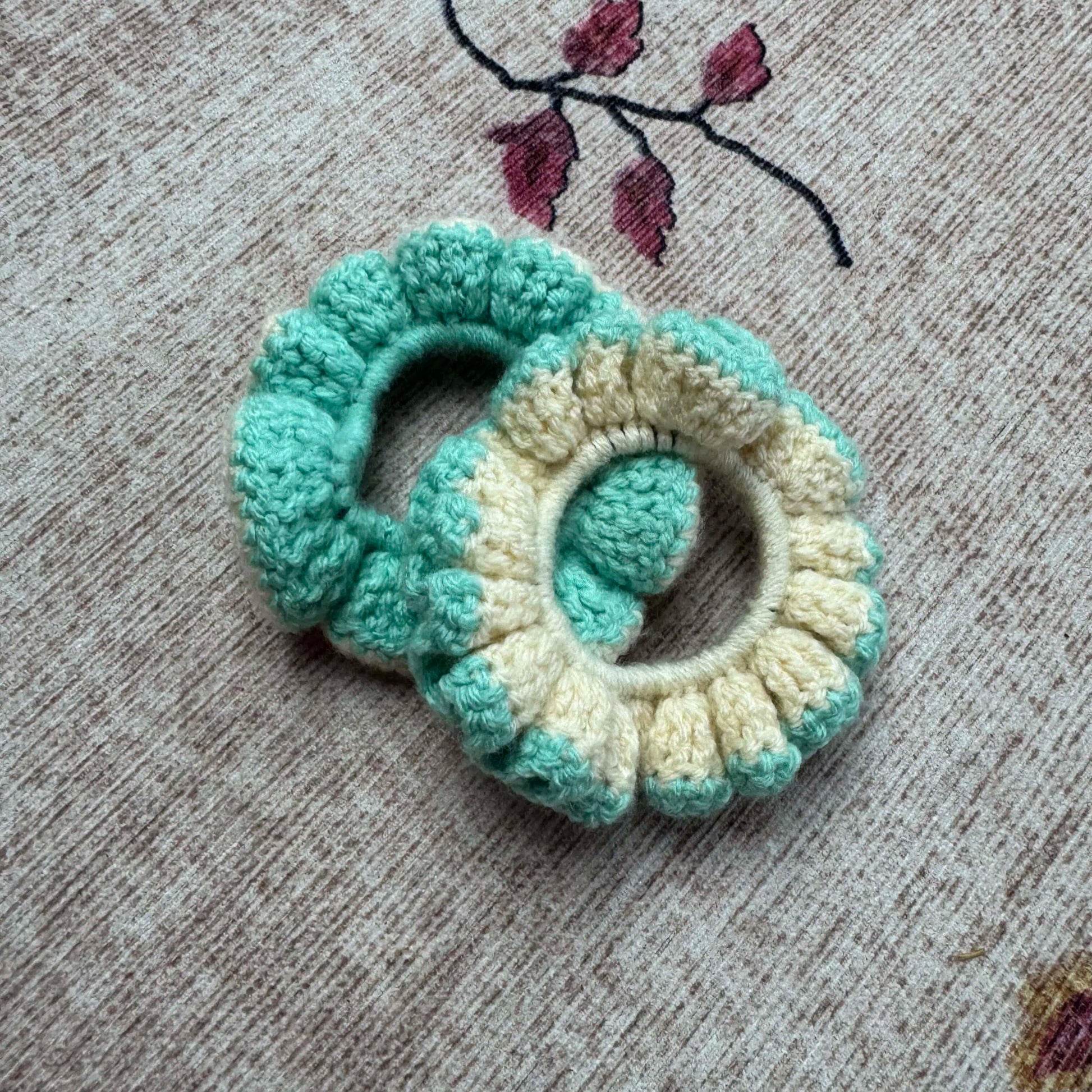 Two crocheted scrunchies in teal and beige on a textured surface.