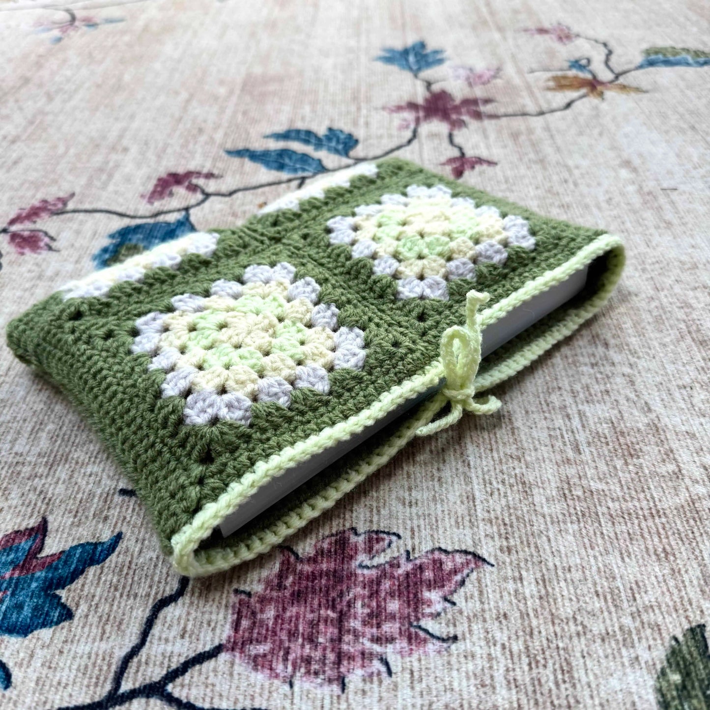 Green crocheted book cover with floral patterns on a floral fabric background
