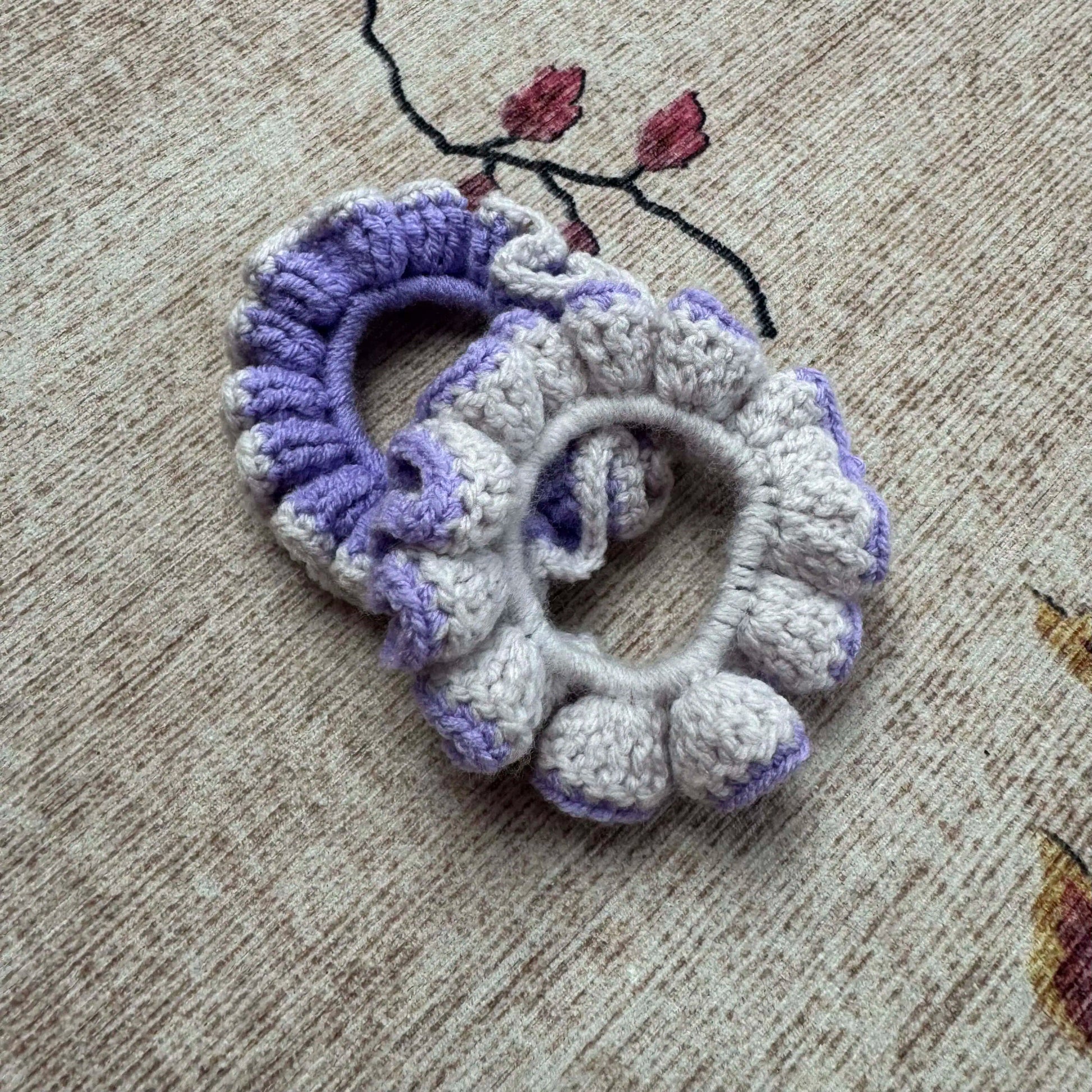 Two crocheted scrunchies, one purple and one white, on a textured surface.