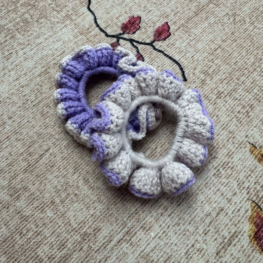 Two crocheted scrunchies, one purple and one white, on a textured surface.