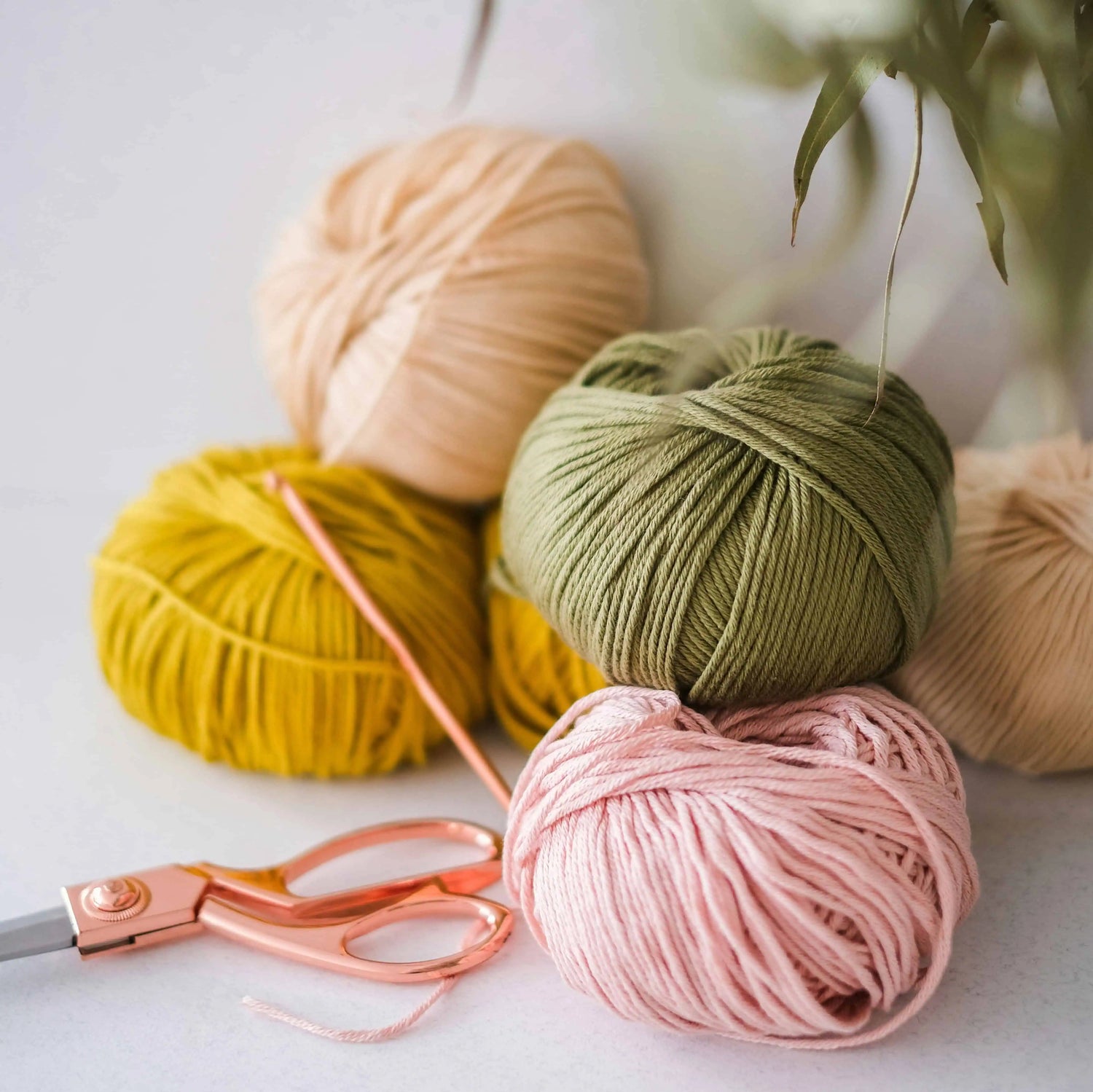 Four balls of yarn in beige, yellow, green, and pink with a pair of scissors on a light background.